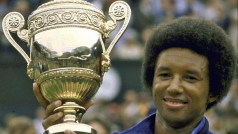 Arthur Ashe, in a blue tracksuit top, smiles as he holds up the Wimbledon trophy after winning the men's final on 5 July 1975.