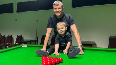 Two-year-old Jude sitting on a snooker table alongside his father