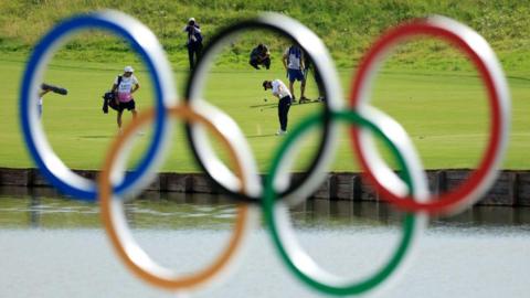 Tommy Fleetwood plays a shot from the fairway at Paris 2024 with the Olympic rings in the foreground 