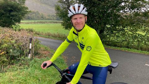 Steve Bate on his black bike, wearing a fluorescent yellow top, cycle helmet and blue cycling trousers with a road junction, hills, grass, trees and mist behind him