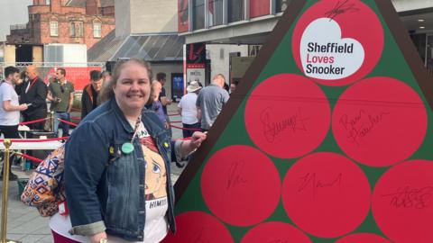 A woman with short brown hair wearing a denim jacket stands outside the Crucible as people queue up to enter for the day. She poses next to a sign that reads 'Sheffield Loves Snooker'