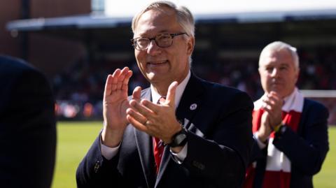 Nick Semaca applauds the Leyton Orient fans, dressed in a dark suit, white shirt and red tie 