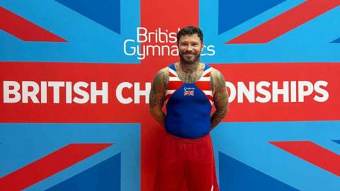 A man wearing a blue, red and white sports vest and shorts with the Union flag as part of the kit. He has short brown hair, a brown beard and tattoos on his arms. He is standing in front of a Union flag banner with "British Gymnastics British Championships" written on it in white.