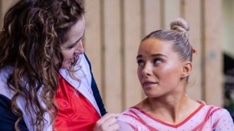 Tracey Skirton-Davies has long brown hair and is looking at Ruby Evans with hair tied back, wearing a white and red top