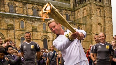England cricketer Paul Collingwood plays a cricket shot with the Olympic torch outside Durham cathedral during the torch relay before London 2012