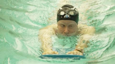 Ela Letton-Jones wears a black swim cap as she swims holding a board