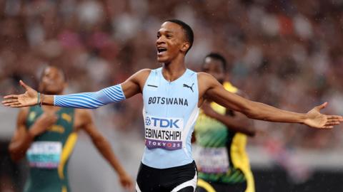 Collen Kebinatshipi seen from hips up, wearing a light blue Botswana running vest with a name bib on the front, black shorts and blue running sleeve covering part of his right arm, stretches out both arms in celebration after winning the 400m final at the World Athletics Championships in Tokyo. Blurred in the background are South African and Jamaican runners trailing in his wake and behind them a large crowd